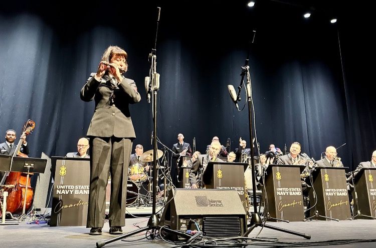 A photo of the U.S. Navy Band Commodores performing Stars and Stripes Forever onstage at the Saenger Theatre, featuring MU1 Amanda Ballantine on piccolo